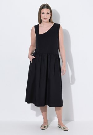 Frau in schwarzem ärmellosem Midikleid mit einem Schulterriemen, Hände in den Taschen, trägt flache Schuhe mit Blumenmuster vor weißem Hintergrund.