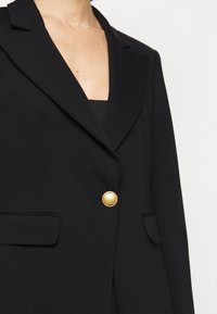 Černý blazer s přizpůsobeným střihem, vybavený jedním zlatým knoflíkem, klopou s vyřezávanými okraji a dvěma předními kapsami. Hladká, strukturovaná tkanina.