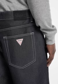 Jeans en denim noir avec un logo en triangle rouge et blanc de Guess sur la poche arrière. Une couture blanche met en valeur le design.