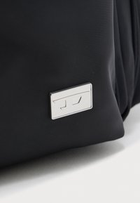 Fekete szövetfelület, az alsó él közelében egy kis téglalap alakú ezüst fém címkével, rajta domborított geometriai mintával.