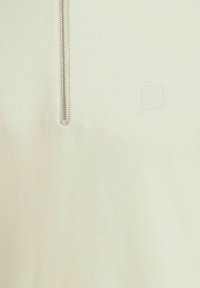 Close-up van crèmekleurige stof met aan de linkerkant een verticale metalen rits en rechts een subtiele reliëfrectangulaire logo.