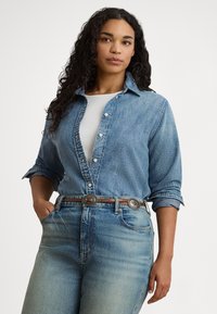 Žena s dlhými kučeravými vlasmi, ktorá má na sebe rozopnutú rifľovú košeľu cez biely top, vyblednuté modré džínsy a hnedý opasok s dekoratívnymi prackami.