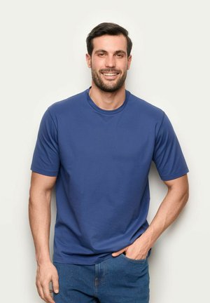 Uomo sorridente con capelli scuri, che indossa una maglietta blu a maniche corte e jeans blu, in piedi con una mano in tasca contro uno sfondo semplice.