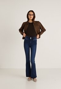 Chaqueta de ante marrón sobre una blusa negra, combinada con vaqueros acampanados de talle alto en azul oscuro. Cuenta con dos botones delanteros y detalles sutiles en los bolsillos.