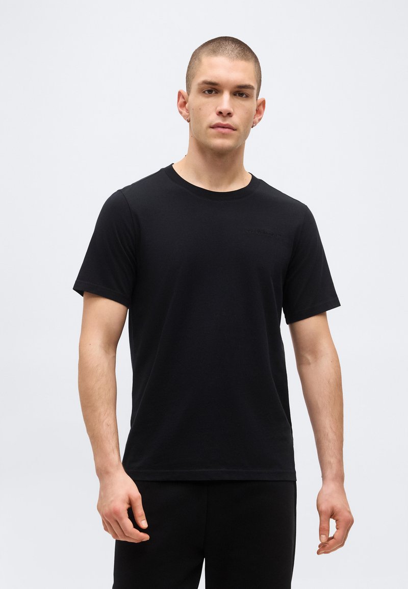 Νέος άνδρας με κοντά μαλλιά, που φοράει απλό μαύρο κοντομάνικο μπλουζάκι και μαύρο παντελόνι, στέκεται μπροστά σε απλό ανοιχτόφωτο φόντο.