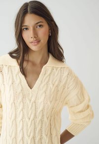 Pullover en maille torsadée couleur crème avec un col en V et un large col. Présente des manches amples et un design texturé. Fabriqué en matière tricotée douce.