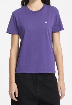 Kobieta nosząca gładki fioletowy t-shirt z krótkim rękawem i małym białym logo na lewej piersi, w połączeniu z czarnymi spodniami.