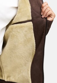 Braune Kunstwildlederjacke mit einem samtigen beigen Futter, ausgestattet mit einem Reißverschluss und einer weichen Textur, die entlang der inneren Kanten sichtbar ist.