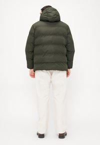Man wearing green hooded puffer jacket, white pants, and brown shoes, standing facing away against plain light background.