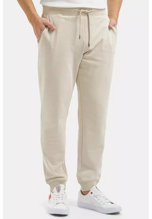 Beżowe spodnie dresowe na sznurku z bocznymi kieszeniami, noszone z białymi niskimi sneakersami na osobie stojącej na jednolitym tle.