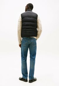 Chaleco de plumas negro sobre un suéter de punto trenzado color crema, combinado con jeans azules y zapatos negros, visto desde la espalda. Diseño sencillo, sin logotipos visibles.