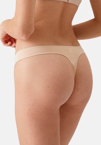 Beige string met een gladde textuur, met een lage taille en minimale bedekking aan de achterkant, die een naadloze afwerking toont.