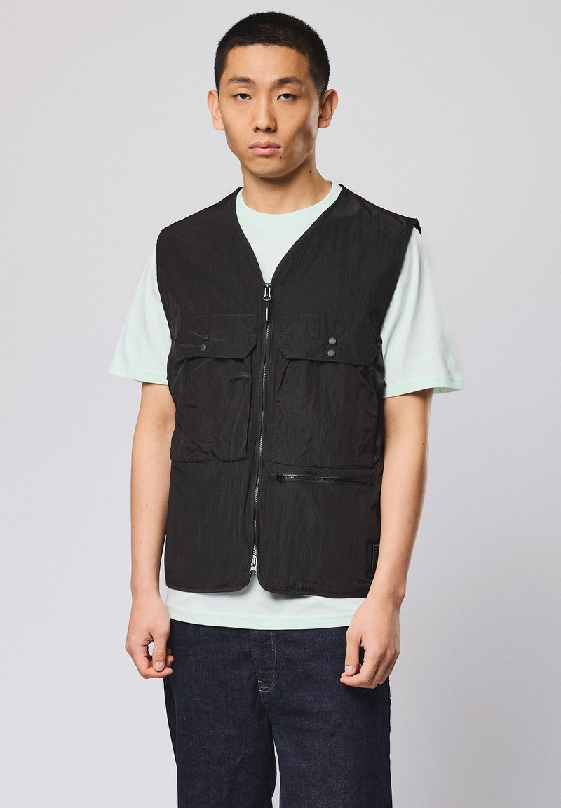Jeune homme aux cheveux courts portant une veste noire zippée avec poches frontales par-dessus une chemise à manches courtes vert clair et un jean sombre, debout devant un fond uni.