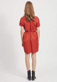 Robe rouge à manches courtes avec une taille cintrée et une jupe ample, en tissu lisse. Portée avec des bottines noires. Vue de dos.