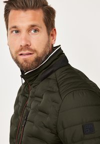 Man with short brown hair and beard wearing a dark green quilted jacket with a zip and high collar looking slightly right.
