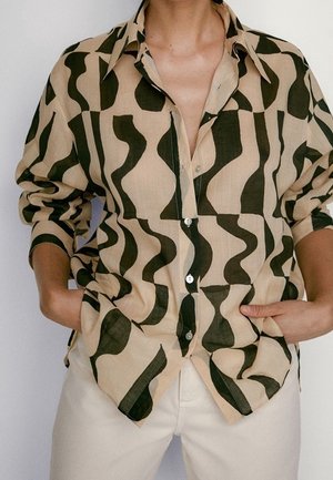 Person wearing beige button-up shirt with dark green abstract wavy patterns, sleeves rolled up, hands in pockets, paired with light-colored pants.