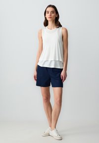 Canottiera bianca a strati sopra pantaloni corti blu navy, abbinata a scarpe da ginnastica bianche. La canottiera ha una consistenza leggera e liscia; i pantaloni corti presentano un design semplice.