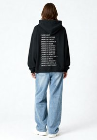 Schwarzer Hoodie mit weißem Text auf dem Rücken, der wiederholte Phrasen auf Französisch zeigt. Kombiniert mit hellblauen, weiten Jeans und weißen Schuhen.