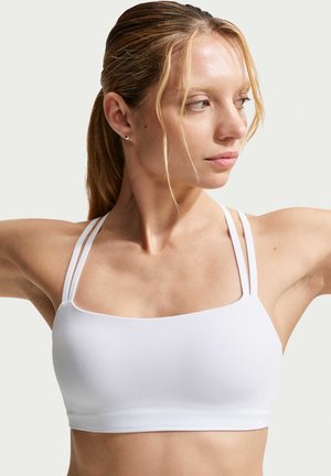 Jeune femme aux cheveux blonds attachés, portant un soutien-gorge de sport blanc avec double bretelles fines, regardant vers sa droite, les bras levés.