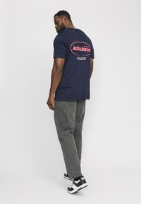 Marineblaues T-Shirt mit rundem rotem Logo auf dem Rücken, graue Hose und schwarz-weiße Sneakers. Einfaches Design, Baumwollstoff, lockere Passform.