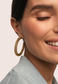 Pendientes de aro dorados con un diseño texturizado y de cuentas. La modelo lleva un top con cuello brillante de color azul claro, sonriente y mostrando los pendientes.
