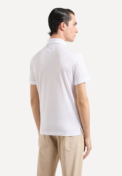 Polo shirt blanc à manches courtes avec un col classique, texture lisse, fentes sur les côtés et subtils détails de couture. Porté avec un pantalon beige.
