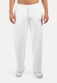 Weiße Sweatpants mit weicher Textur, elastischem Bund, Seitentaschen und einem kleinen goldenen "Smilest"-Logo auf dem linken Oberschenkel.