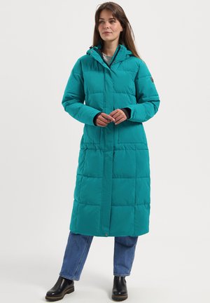 Woman wearing a long teal quilted winter coat with hood, paired with blue jeans and black ankle boots, standing against a plain background.