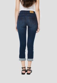 Sötétkék, rövidített farmernadrág, testhez simuló fazonnal, felhajtott szegéllyel, két hátsó zsebbel és bőrből készült címkével a derékrészen.