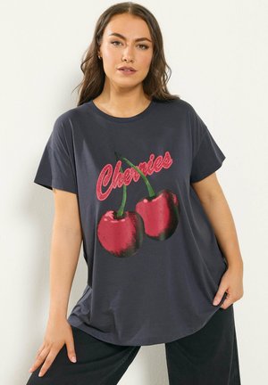 Młoda kobieta w luźnej ciemnoszarej koszulce z czerwonymi wiśniami i napisem "Cherries", pozująca na jasnym tle.