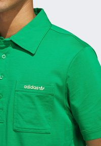 Grön polo skjorta i bomull, med klassisk krage, framficka och broderad adidas-logotyp i guld. Slät textur.