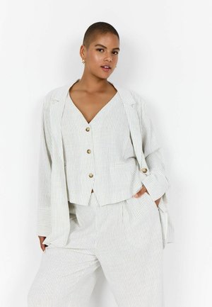 Vrouw met kort haar die een wit pak met gouden knopen en fijne strepen draagt, staand met één hand in de zak tegen een effen achtergrond.