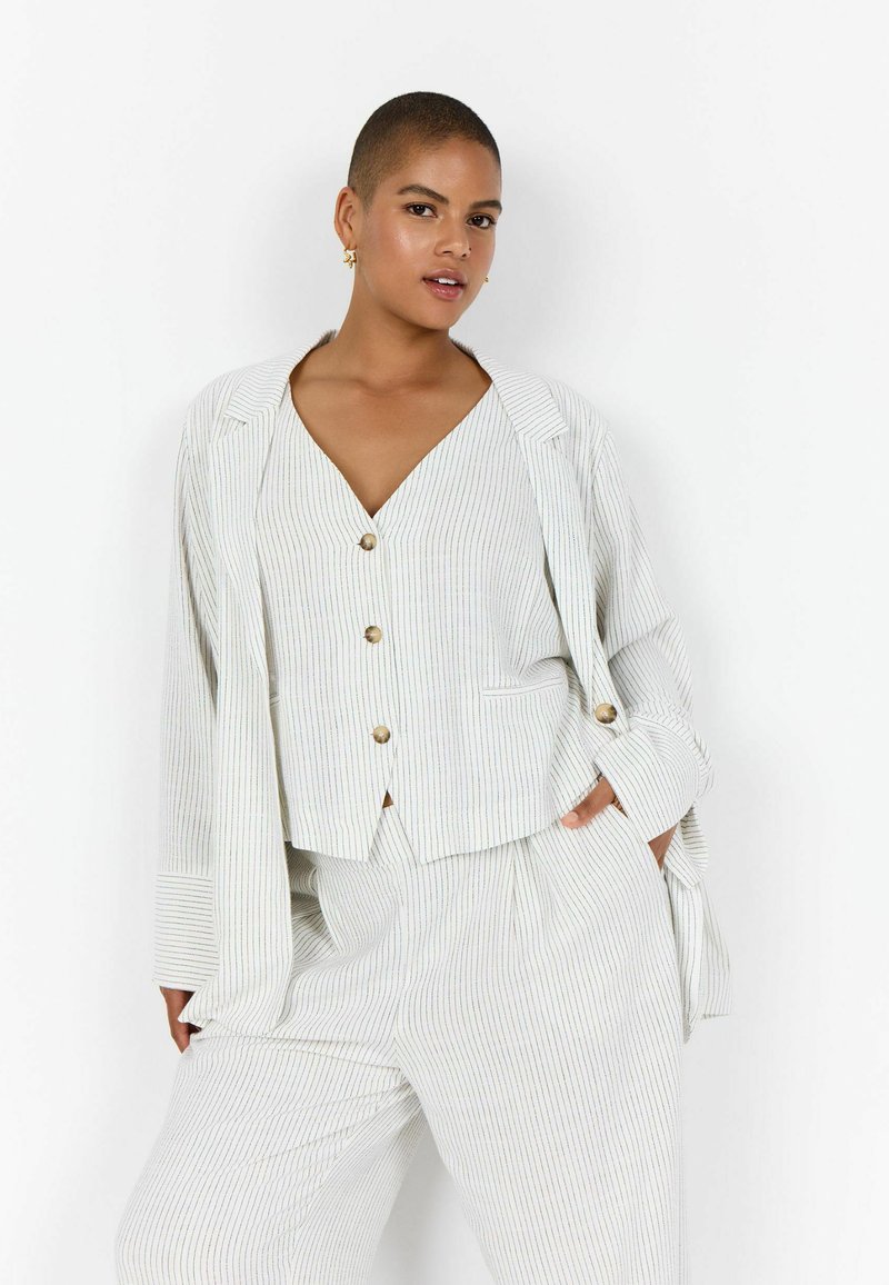 Rövid hajú nő fehér, arany gombos csíkos öltönyben, egyik kezét a zsebében tartva, egyszínű háttér előtt áll.