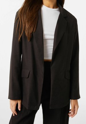 Vrouw draagt een zwart oversized blazer over een witte cropped top, met losse zwarte broek, staand tegen een effen witte achtergrond.