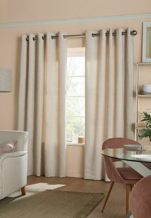 Des rideaux beiges pendent d'une tringle en métal au-dessus d'une fenêtre dans un salon cosy avec un canapé beige, une table à manger en verre et des chaises roses douces.