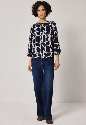 Kvinde iført en langærmet top med sort-hvidt geometrisk mønster, mørkeblå jeans og brune sko, stående mod en ensfarvet baggrund.