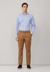 Camisa de botones a rayas azul claro y blanca, combinada con pantalones chinos marrones y zapatos oscuros sin cordones, de pie contra un fondo neutro.