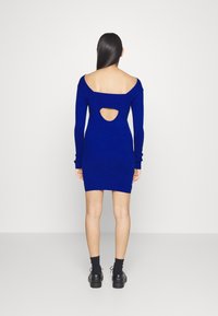 Modrá pletená šaty s dlouhými rukávy, s otevřeným výřezem na zádech a přiléhavým střihem, doplněné černými botami a ponožkami.