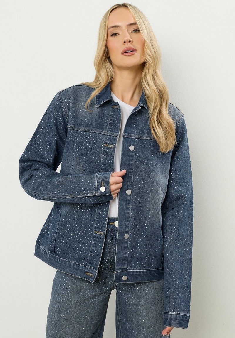 Denimjack met een donkerblauwe kleur en een licht verouderde uitstraling. Versierd met zilveren studs over het geheel. Voorzien van een knoopsluiting en zakken.