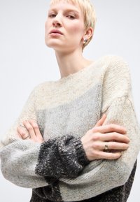 Άτομο με κοντά ξανθά μαλλιά που φορά ένα πλεκτό πουλόβερ με υφή και πολύχρωμους χρωματικούς συνδυασμούς και ασημένια σκουλαρίκια κρίκους, με τα χέρια σταυρωμένα με αυτοπεποίθηση.