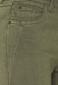 Olivově zelená denimová látka s kapsou na zadní straně, která má detailní prošívání a černé kovové akcenty. Texturovaný povrch s jemným leskem.