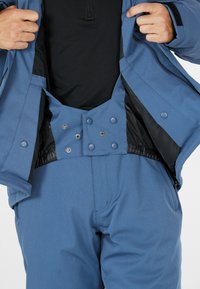 Hände, die die inneren Druckknöpfe einer blauen Outdoorjacke und der passenden Hose über einem schwarzen Shirt verstellen.