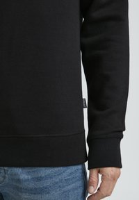 Black sweatshirt with ribbed cuffs and hem, featuring a small tag on the side. The material appears soft and textured with a fitted design.