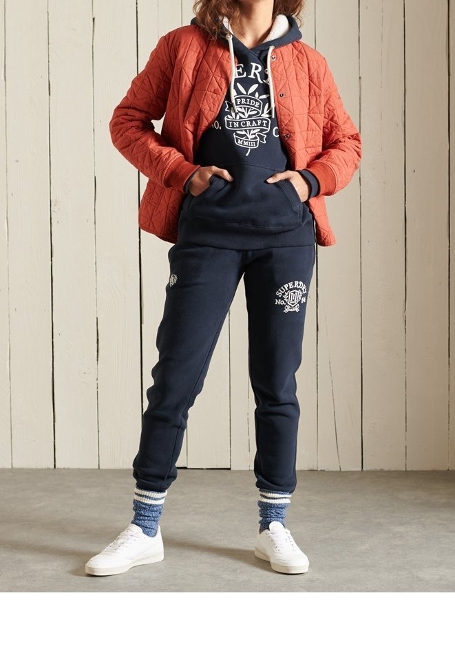Personne portant un sweat à capuche bleu marine avec logo et un pantalon de survêtement, une veste matelassée orange, des chaussettes rayées bleues et des baskets blanches, debout à l'intérieur.