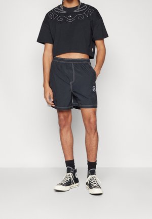 Sort cropped skjorte med hvidt grafisk design, kombineret med mørke shorts med hvide syninger og et logo. Sorte høje sneakers fuldender looket.