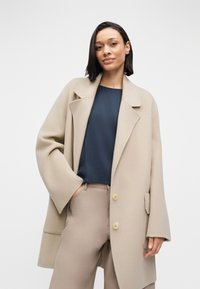 Abrigo de lana beige oversize con solapas anchas y dos botones, superpuesto sobre una blusa azul oscuro y pantalones beige. Tela texturizada, ajuste casual.