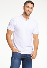 Valkoinen puuvillapaita, jossa on klassinen kaulus, lyhyet hihat ja pieni musta logo rinnassa. Yhdistetään sinisiin farkkuihin.