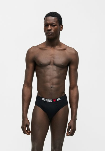 Άνδρας χωρίς πουκάμισο με σκουρόχρωμο δέρμα που φοράει μαύρα εσώρουχα Moschino με κόκκινο σύμβολο καρδιάς και κείμενο "YOU" στη μέση, στέκεται μπροστά σε απλό φόντο.