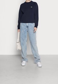 Sweatshirt bleu marine avec manches raglan et logo vert, associé à un jean en denim bleu clair oversize. Sac à main tissé blanc et baskets blanches.