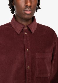 Camisa de pana burdeos con cierre de botones en la parte delantera, cuello puntiagudo, bolsillo en el pecho y tela texturizada. Presenta botones marrones y detalles cosidos.